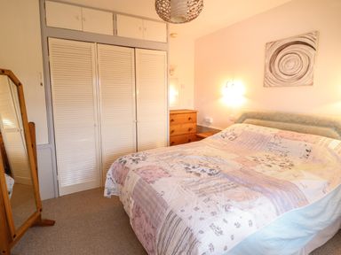 A bedroom with a bed covered with a patterned quilt a wooden dresser a floor mirror and white wardrobes at Morlyn Guest House Apartment in Llandanwg near Harlech