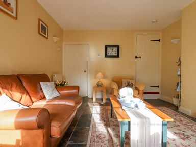 A living room with a brown leather sofa an armchair a wooden coffee table with a table runner and tea set a wall clock and doors at Bodkin Cottage in Dunster