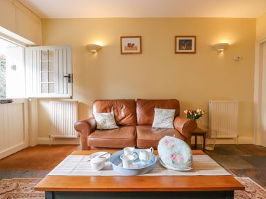 A living room with a brown leather sofa two cushions a wooden coffee table with tea set and a floral tea cozy Bodkin Cottage Dunster