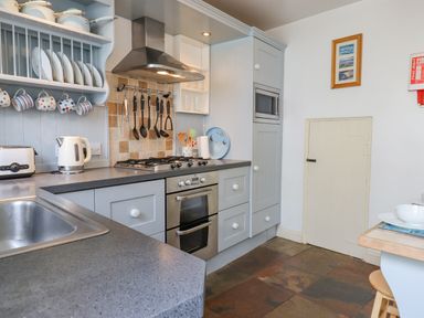 A kitchen with a stainless steel oven stove a sink a kettle a toaster hanging mugs and cooking utensils at Bodkin Cottage in Dunster