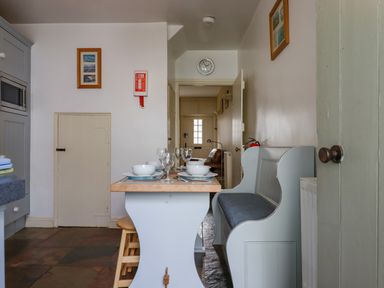 A dining area with a wooden table set with bowls and glasses a bench and stools a clock on the wall and a doorway in Bodkin Cottage in Dunster