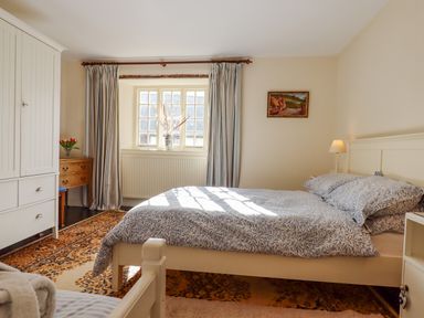 A bedroom with a bed, wardrobe, wooden dresser, window with curtains, lamp, and framed picture at Bodkin Cottage in Dunster