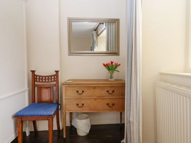 A wooden chair with blue cushion next to a wooden chest of drawers with red flowers in a vase and a mirror above at Bodkin Cottage in Dunster