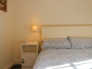 A bedroom with a white headboard bed with patterned bedding and a white nightstand with a lamp and a clock at Bodkin Cottage in Dunster