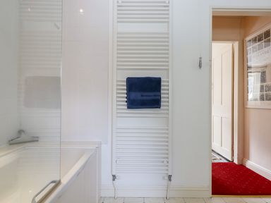 A bathroom with a white bathtub white tiled walls a white towel radiator with a blue towel and an open door showing a hallway with a red carpet at Bodkin Cottage in Dunster