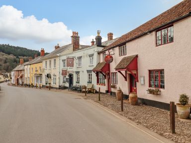 A street with a row of connected buildings including a pub and restaurant with outdoor seating at Bodkin Cottage in Dunster