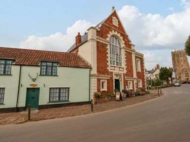 A street with a pale green house with a tiled roof and a large brick building with arched windows and a bench outside in Dunster