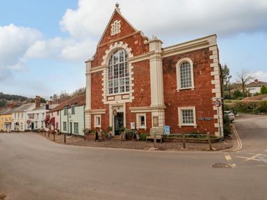 A street corner with a large brick building and smaller colorful houses along the road at Bodkin Cottage in Dunster