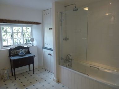 A bathroom with a bathtub and shower combination a small wooden cabinet with a ceramic pitcher and bowl a window with multiple panes and cabinetry on the side at Bodkin Cottage in Dunster