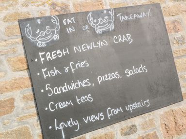 A chalkboard menu on a stone wall listing fresh Newlyn crab fish and fries sandwiches pizzas salads cream teas and lovely views from upstairs