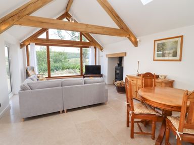 A living room with wooden beams a gray sofa a round wooden dining table and large window at The Nook in Jacks Green near Painswick