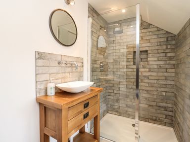 A bathroom with a wooden vanity with a white vessel sink and a round mirror above it and a walk-in shower with a glass partition and brick-style tiled walls at The Nook in Jack's Green near Painswick