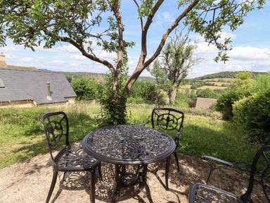A garden patio with a metal table and chairs under a tree overlooking fields and a house at The Nook in Jacks Green near Painswick