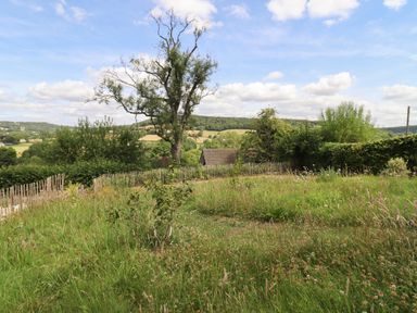 A grassy outdoor area with a wooden fence and trees overlooking fields and hills at The Nook in Jack's Green near Painswick