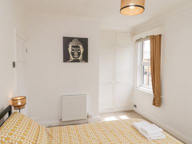 A bedroom with a bed covered in a yellow and white patterned duvet a lamp a window with brown curtains and built in white cupboards at 89 Caledonia Street Scarborough