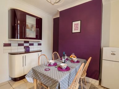A dining area with a table set for four with bowls wine glasses and a bottle on a polka dot tablecloth next to a purple accent wall and white fridge at 89 Caledonia Street Scarborough