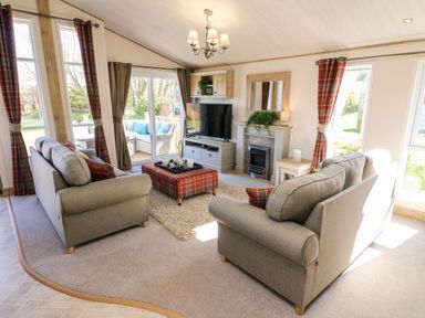 A living room with two sofas a television on a cabinet a fireplace and plaid curtains at Curlew Lodge in Carnforth