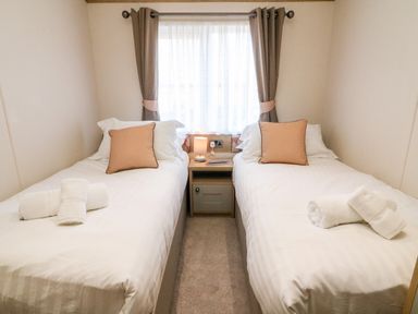 A bedroom with two single beds separated by a nightstand with a lamp and window with curtains at Curlew Lodge in Carnforth