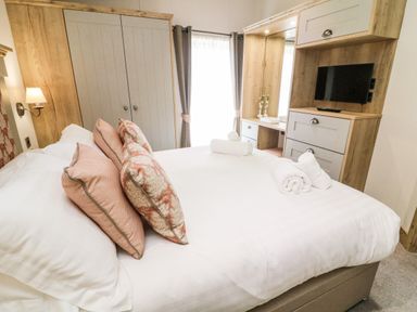 A bedroom with a bed with pillows and rolled towels a wardrobe and wooden furniture with a mounted tv at Curlew Lodge in Carnforth