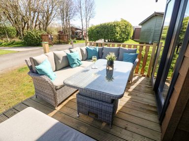 An outdoor patio with a wicker corner sofa with cushions around a glass top table and a potted plant at Curlew Lodge in Carnforth