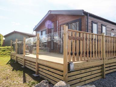 An exterior view of a wooden lodge with glass panel railings and outdoor seating at Curlew Lodge in Carnforth