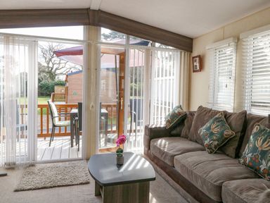 A living room with a sofa a coffee table and glass doors opening to a deck with chairs and an umbrella at Audrey's Lodge Foxburrow Hang in Fritton