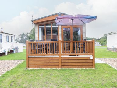 A small wooden deck with chairs and a table under an umbrella outside a cabin at Audreys Lodge Foxburrow Hang in Fritton