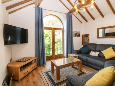 A living room with wooden beams, a large arched window with blue curtains, a wall-mounted TV, a wooden coffee table, and a gray sofa with cushions at Ty Gwennol in Llanddona near Beaumaris