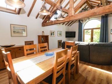 A dining area with a wooden table and chairs and a living room with a gray sofa and a mounted TV at Ty Gwennol in Llanddona near Beaumaris