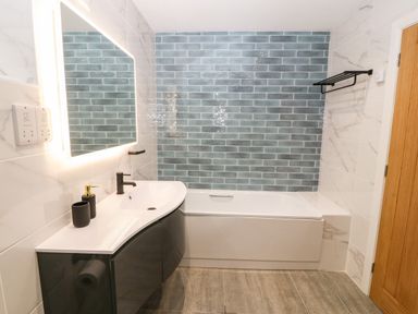 A bathroom with a bathtub, a curved sink with a faucet, a backlit mirror, tiled walls, and a wooden door at Ty Gwennol in Llanddona near Beaumaris