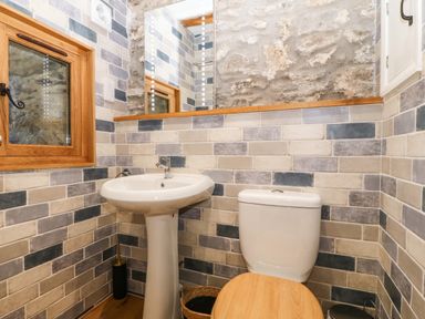 A bathroom with a pedestal sink a toilet with a wooden seat a window and a large mirror with tiled walls and stone accent at Ty Gwennol in Llanddona near Beaumaris