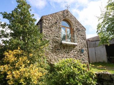 A stone building with a large arched window and surrounding green and yellow bushes at Ty Gwennol in Llanddona near Beaumaris