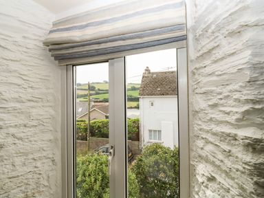 A window with striped blinds between textured white walls showing houses and greenery outside at Alexanders Cottage in Frogmore