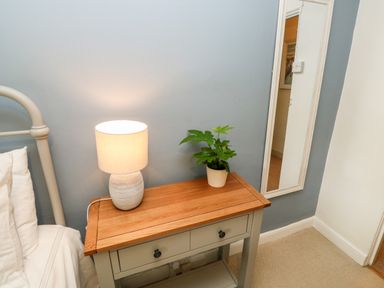 A bedside table with a lamp and a potted plant next to a bed and a wall mirror at Alexanders Cottage in Frogmore