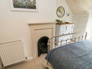 A bedroom corner with a metal bed frame covered with a blue blanket a small fireplace a white radiator a white dresser with plants and a round mirror above at Alexanders Cottage in Frogmore