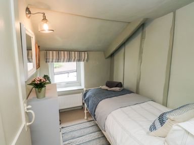 A small bedroom with a single bed a striped window blind a white cabinet with a flower pot and a wall light at Alexanders Cottage in Frogmore