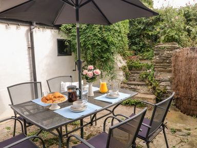 An outdoor dining area with a metal table and chairs set with pastries coffee and orange juice under a large umbrella at Alexanders Cottage in Frogmore