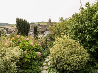 A garden with stone path surrounded by green shrubs and flowering plants with houses and hills in the background at Alexanders Cottage in Frogmore