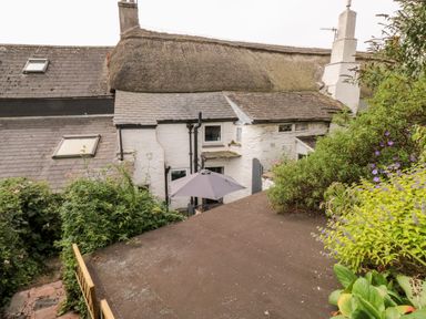 Exterior view of a thatched roof cottage with white walls, garden plants, and an outdoor umbrella at Alexanders Cottage in Frogmore