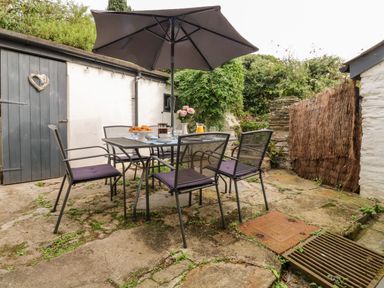 An outdoor patio with a table and four chairs under a large umbrella and a wall with a door and greenery at Alexanders Cottage in Frogmore