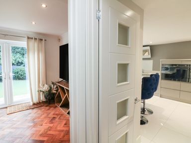 A room with wooden parquet flooring and a TV next to a kitchen area with blue bar stools and tiled floor at 4 Woodland Park in Northam