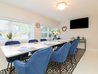 A dining room with a long white table surrounded by blue chairs and a wall mounted television at 4 Woodland Park in Northam