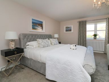 A bedroom with a grey upholstered bed a white quilt bedside tables with lamps and framed pictures on the walls at 4 Woodland Park in Northam