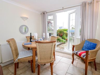 A dining area with a round wooden table and wicker chairs next to open glass doors leading to a garden at Angorfa Bach in Abersoch