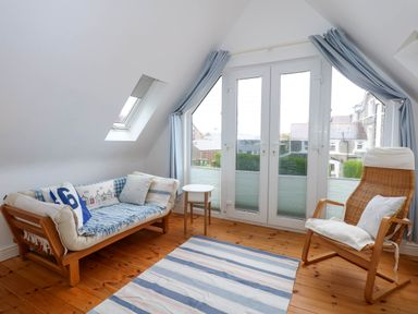A small attic room with a wooden futon sofa with cushions a round side table a wooden armchair with cushions striped rug and glass doors with curtains at Angorfa Bach in Abersoch