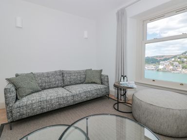 A sitting area with a grey patterned sofa two cushions round ottoman side table with binoculars and magazine window with a view at Upper Tremorvah in Dartmouth