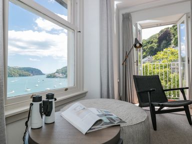 A room with a chair lamp and table with binoculars and magazine near windows overlooking water and trees at Upper Tremorvah in Dartmouth
