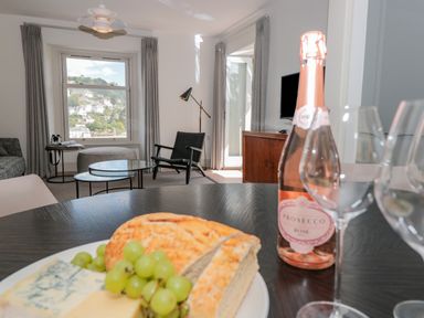 A dining table with bread grapes cheese and a bottle of prosecco in a living room with a window and armchair at Upper Tremorvah in Dartmouth