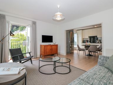 A living room with a sofa a black chair a wooden cabinet and a glass coffee table opening to a kitchen with a dining table at Upper Tremorvah in Dartmouth