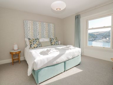 A bedroom with a double bed cushions and bedside tables with lamps next to a window showing a water view at Upper Tremorvah in Dartmouth